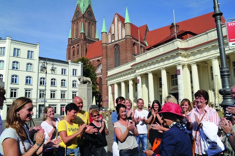Eine Gruppe Touristen in sommerlicher Kleidung steht klatschend vor einer Touristenführerin mit pinkem Hut. Die Gruppe befindet sich auf dem Marktplatz, im Hintergrund ist der Dom. 
