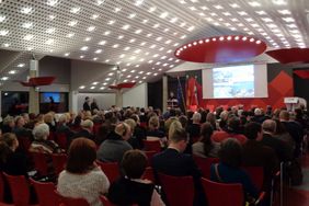 Ein Publikum auf roten Stühlen sitzt vor einer Bühne mit einer Power-Point-Präsentation. Beate Schlupp steht auf der Bühne an einem Podium neben den Flaggen Eurpoas, Deutschlands und Mecklenburg-Vorpommerns.