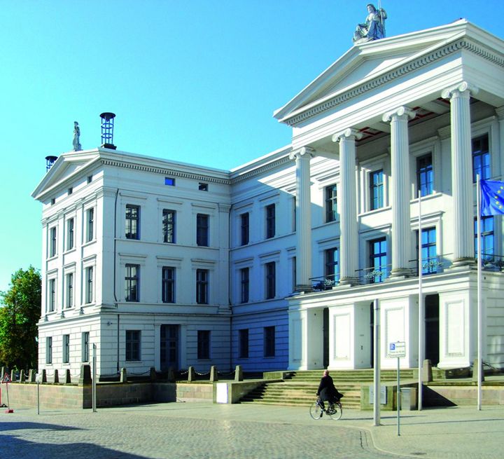Eine Aufnahme des äußeren Flügels des Kollegiengebäude 1 und die Säulenhalle über der Terrasse auf einem umlaufenden Sockel. Eine Europaflagge weht vor dem Gebäude an einem Fahnenmast.