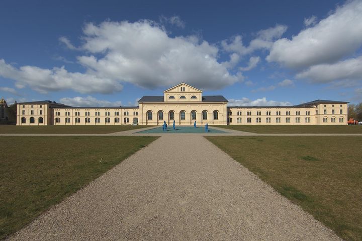 In der Rückansicht ist der Marstall im klassischen Stil von zwei zweigeschossigen Eckpavillons begrenzt. Auf dem Hof vor dem länglichen Gebäude befindet sich eine Installation mit blauen Pferdefiguren. 